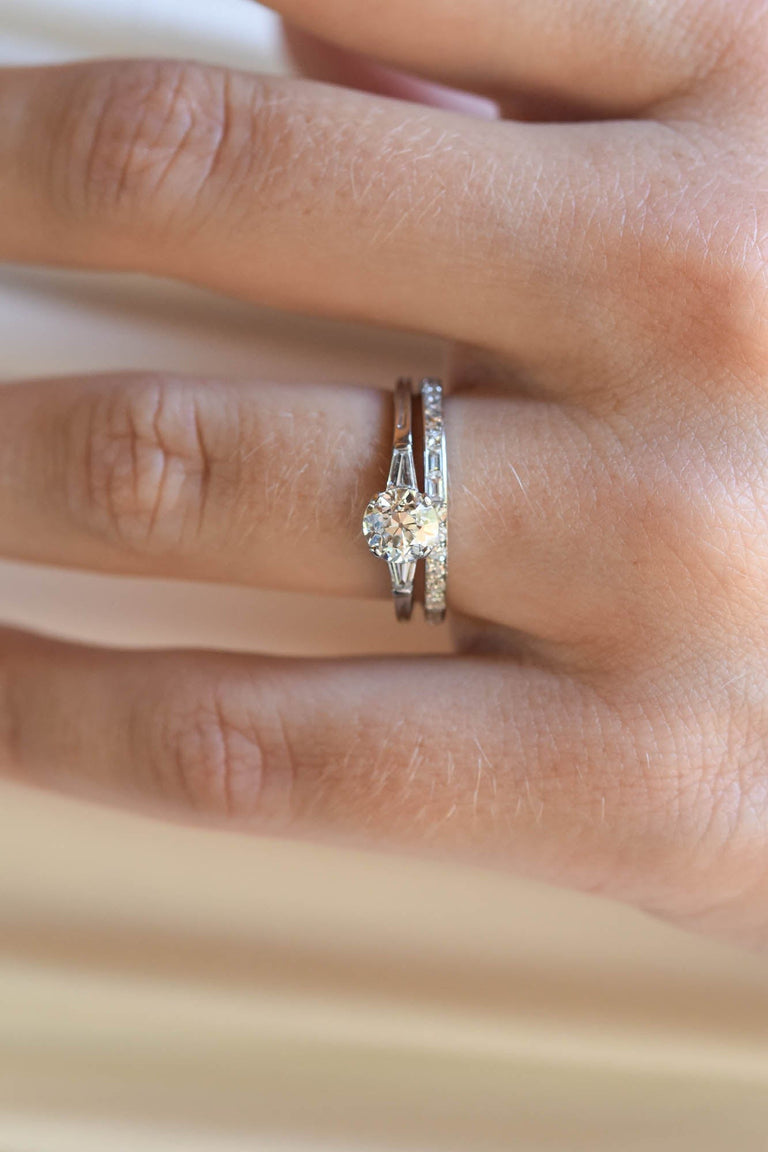 Hand wearing a platinum vintage inspired wedding band and engagement ring on a light background